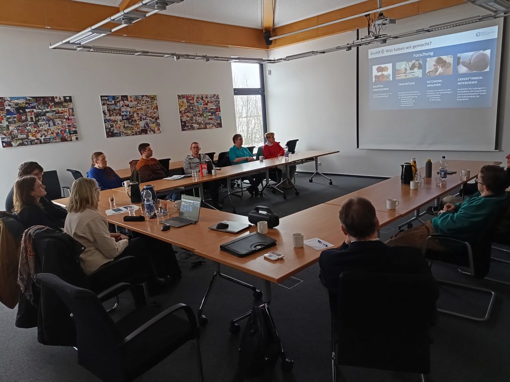 Es sitzen Menschen in einem Besprechungsraum an Tischen und schauen auf eine Leinwand mit einer Präsentation.