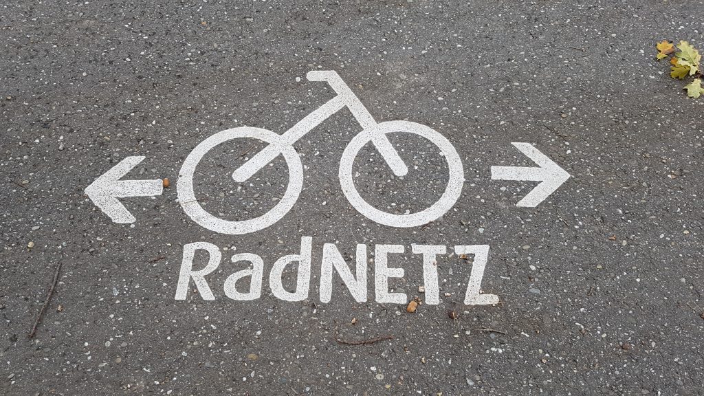 Das Foto zeigt einen Boden auf Asphalt, der die Aufschrift "RadNETZ" sowie ein Piktogramm mit einem Fahrrad und zwei Pfeilen zeigt.